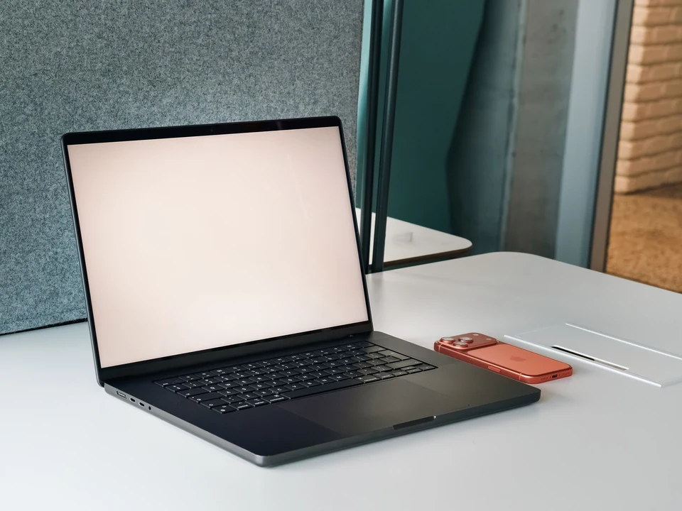 Black laptop with a blank white screen on a clean desk beside a phone and bright window.